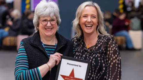 LNER Lesley on the left is smiling and holding up a certificate in a black frame with a star on it honouring her mother, Gladys. her daughter, Vicky, stands beside her also smiling