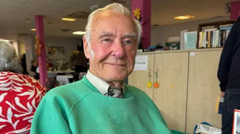 Ted has white hair and prominent sideburns and is sitting at a table inside a community centre he's wearing a bright green sweatshirt over a collared shirt and patterned tie.