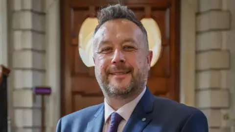 Walsall Council A man wearing a suit smiles at the camera. He has short, brushed-back gark hair, and a slightly greying beard. 