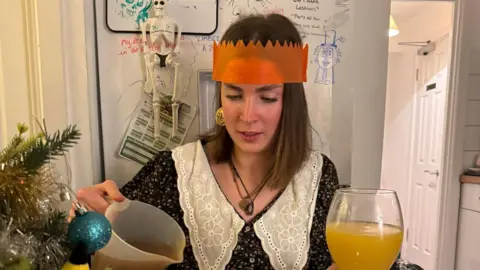 Izzy White A woman sat in a student kitchen at Christmas time wearing an orange party hat and pouring gravy onto a roast dinner. 