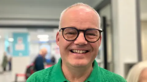 A man with short grey hair smiles at the camera. He wears big round black glasses and a green polo shirt