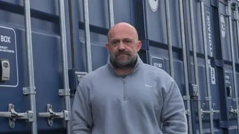 Richard Thomas A bald man with a stern expression looking towards the camera, he's wearing a light blue jumper and black jeans. He stands in front of blue storage containers.