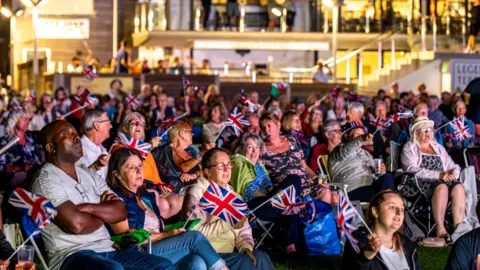 A photo which shows a crowd of people all sitting down. There are about 50 people in the photo. Most are holding union Jack flags.