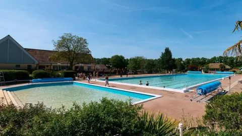 Two outdoor pools at Hitchin Lido. There are bushes in the foreground. There is blue sky above.