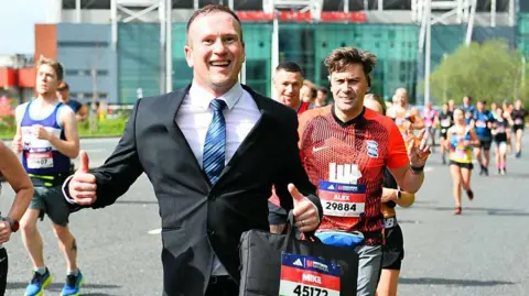 Mike Jones running in the London Marathon in 2024 wearing a black suit and carrying a briefcase, with runners behind and to the side of him.