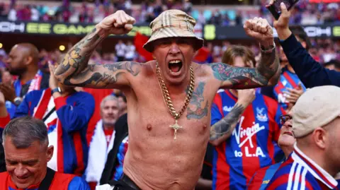 Reuters A shirtless Crystal Palace fan celebrates in the stands after winning the FA Cup. He wears a gold chain around his next with a crucifix and a bucket hat.