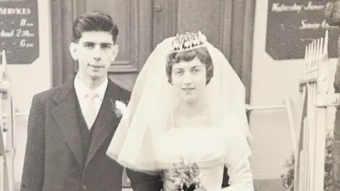 David Bishop An old image of John and his wife on their wedding day. John, on the left, wears a black and white suit and has dark black hair. His wife, on the right, wears a white dress and a large vale with a silver tiara. 
