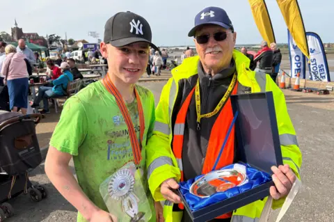 Jodie Halford/BBC Albert in a green T-shirt and black baseball cap, with a medal ribbon round his neck. He has mud splatters on his T-shirt. To his left is Brian Farrington, an older man in sunglasses and a baseball cap, holding a trophy or silver plate.