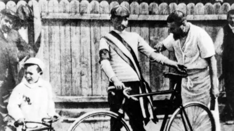 Getty Images A black and white image of Maurice Garin. He is standing behind his bike as another man inspects his arm. A small child can also be seen to their left trying to hold up another bike,