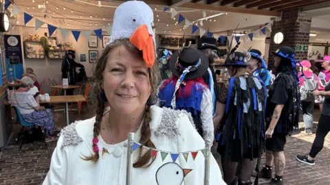 A woman with brown hair in plaits is dressed like a seagull with a white paper and orange beak on her head. Behind her is a group of morris dancers wearing all black.