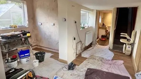 Martin Giles/BBC Dust sheets are on the floor of the Shortt's kitchen area, which has been stripped of all units back to plaster. A work bench is to the left hand side, surrounded by buckets and various building materials.