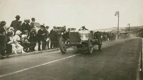 Autocar Magazine Crowds, as far as the eye can see, press against a rope barrier as a Talbot racing car speeds by. A lady dressed entirely in white, from stockings to hat, leans over to get an even closer view