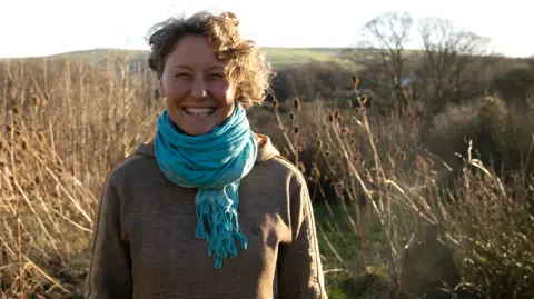 Rosie is standing in the grounds of Brithdir Mawr surrounded by grass and bushes with hillside behind her.  She has short curly fair hair and is wearing a brown long-sleeved top and a blue cotton scarf. 