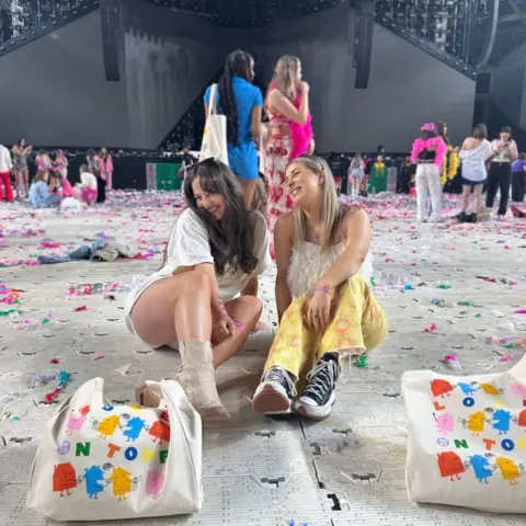 Georgia Prickett Two girls sat on the floor, surrounded by multi-colour streamers and patterned tote bags, smiling and laughing. They are at a concert venue and in the background are groups of other people