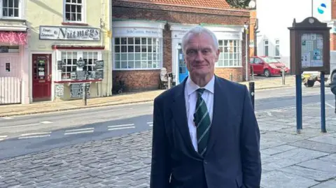 MP Graham Stuart standing in the partially cobbled street of a market town. He is wearing a blue suit and a green tie and is looking at the camera. Behind him there is a notice board and a number of old-fashioned-looking shops.