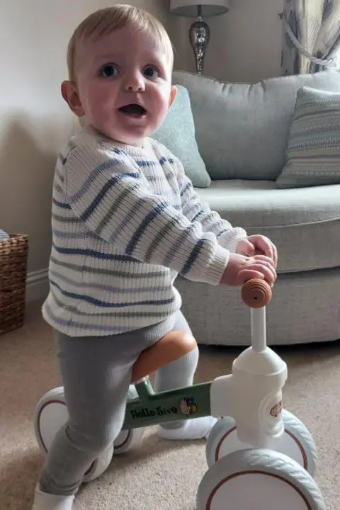 Family handout Finley - a toddler with some blond hair on his forehead - is sitting on a children's pushbike wearing a white jumper with blue and green stripes and grey trousers. He has his mouth open looking to the side, next to a small grey sofa.