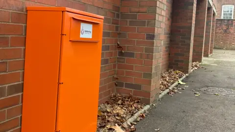Kettering Town Centre A tall orange box in front of a brick wall next to a path.