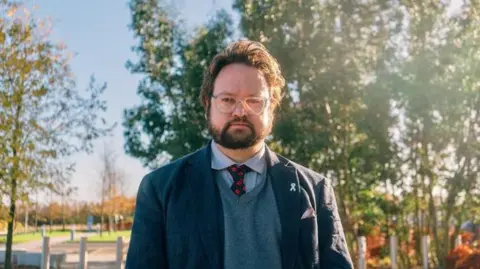 Head and shoulder shot of a man with slightly unkempt dark hair and a beard. He is wearing a dark blue jacket over a blue v-neck jumper and light blue shirt with a red and blue tie, and spectacles with light frames. He is standing in a park or open area with trees behind him.