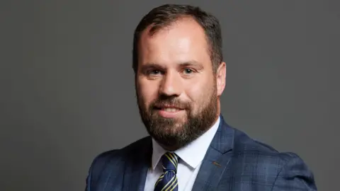 Sean Woodcock's parliamentary photo portrait. He has short brown hair and a  brown beard. He wears a blue suit, blue and gold striped tie, and white shirt.