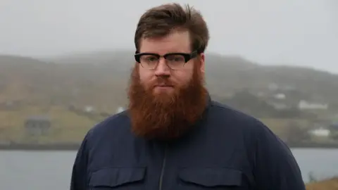 The councillor wears glasses and has a large beard. He is standing outside and in the background is a loch and beyond that a hillside dotted with houses.