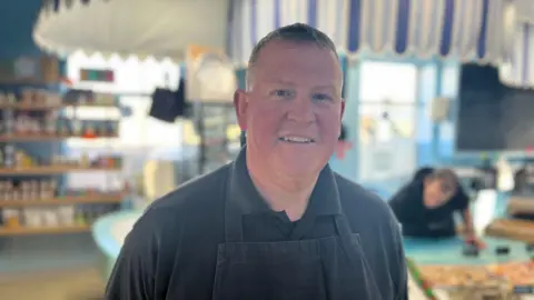 Jason Hamon in his shop, he is wearing an apron and a dark grey polo shirt. He has short dark hair.