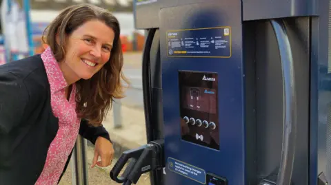 Councillor Natasha Bradshaw is smiling at the camera as she leans towards the new charger. The charger consists of an electric plug-in, a dial and buttons. The councillor is wearing a pink blouse beneath a dark jacket and she has long dark hair.  