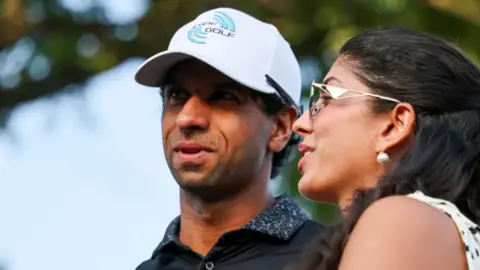 Getty Images Rai is wearing a white cap and navy top and his wife had a summer blouse on with her hair tied back and sunglasses