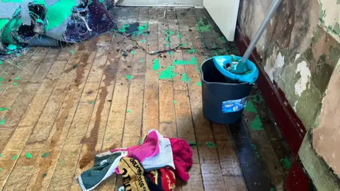 Elizabeth Baines/BBC A hardwood floor with a mop and bucket. Carpet has been ripped up and towels are soaked on the floor.