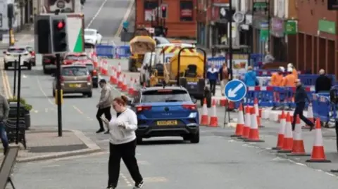 The high street in Ashton-in-Makerfield with pedestrians and roadworks.