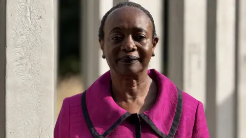 Thelma Stober is seen wearing a bright pink jacket with black trim stands in front of tall pillars at the 7/7 memorial. She is looking directly at the camera.
