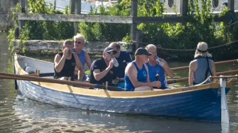 PA/Ben Birchall Seven middle-aged rowers in a large wooden rowing boat are pausing to drink in the middle of a river. There is a larger boat anchored in the background behind a large wooden fence and greenery
