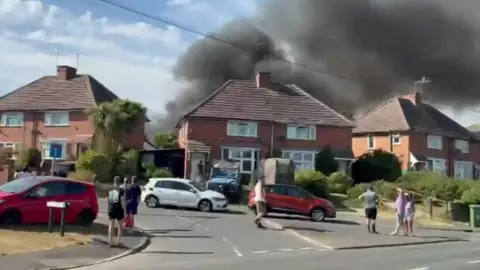 James Lees People stand in the street as smoke billows behind some detached houses. 