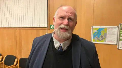 Amy Holmes/BBC Tom Wootton is standing in a wood-panelled room that appears to be an office or meeting space. He is dressed in a dark blue suit jacket over a black sweater, with a white collared shirt and patterned tie. Behind him are wall-mounted evacuation diagrams and a row of black cushioned chairs with wooden frames.