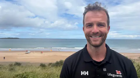 Michael Thompson stands on high ground overlooking White Rocks beach. He has short brown hair and a short brown beard and is wearing a dark polo shirt with the RNLI lifeguard logo.