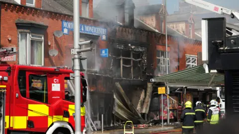 The shop front is gutted and black with smoke coming out of the premises. A fire engine is parked nearby. There are three firefighters on the street.