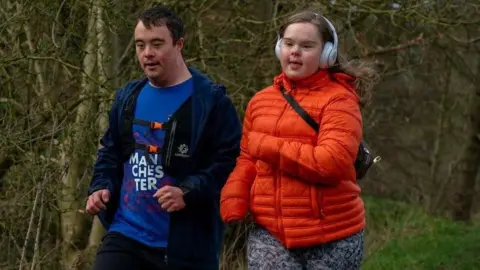 Jillian Perry A man with a blue T-shirt, hooded top and running vest is running alongside a woman wearing an orange padded jacket and leggings.