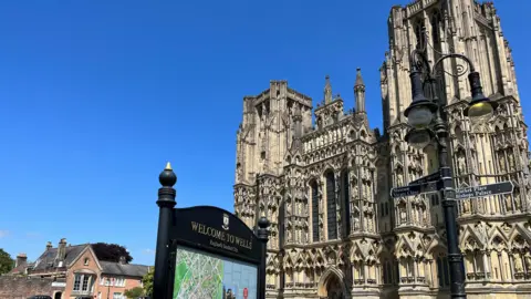 Wells Cathedral is on the right, behind a sign saying Welcome to Wells. The sky is blue.