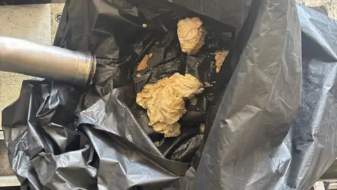 A metal pipe rests on top of a black rubbish bag containing large beige clumps of refuse.