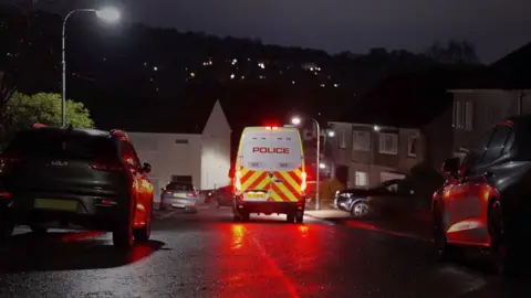 BBC News The rear of a police van, with red lights glowing off a wet road surface middle of shot, flanked by cars and houses on each side.