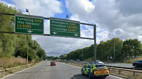 A general view of the A45 near Northampton. Cars can be seen travelling down both sides of the road including a police car. A car sign can be seen overhanging the carriageway.