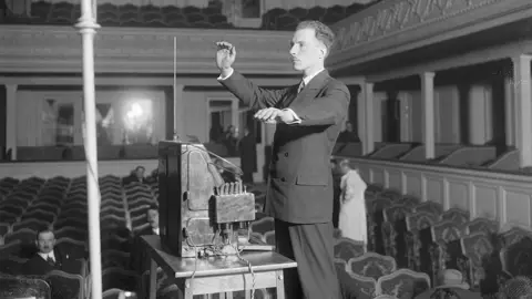Getty Images Leon Theremin playing a Theremin in an theatre, which is largely empty apart from a couple of people sitting in the seats. The inventor's musical instrument is on a small table in front of him, and his hands are positioned about the box in mid-air as he plays it.