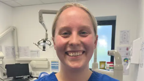 A young woman is wearing blue dental scrubs and has a University of Plymouth name badge on that says "Isabelle Adams, dental student". She is smiling.