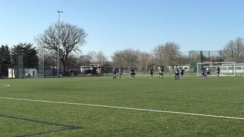 A shot of an artificial football pitch with about a dozen players in the distance. It is surrounded by high fencing, trees, and more open space.