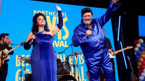 ITV Cymru Wales / BBC Cymru Wales Jones and Speirs performing on stage dressed in electric blue costumes as Priscilla and Elvis. Behind them is the Porthcawl Elvis Festival blue branding and a band performing. Jones, on the left, wears a long silk dress, silk arm covers and has a dark brown wig on. Speirs, is stood next to her, wearing a long sleeve silk shirt and silk trousers. 