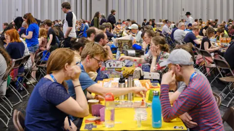 A large hall with rows of people sitting around yellow tables playing board games