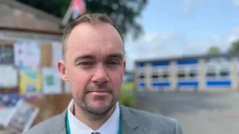 A man with short brown hair, stubble, a white shirt, grey suit jacket and green lanyard is standing in a playground. The playground behind him is empty and blurred from vision.
