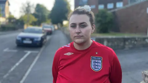 A woman in a red England football shirt stands on a street with a red brick block of flats behind her. She has her hair tied up and is looking directly at the camera.
