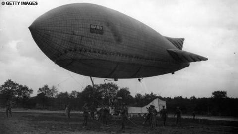 Black and white image of an Airship floating slightly above the ground, with a light aircraft and ground crew below it. A small label on the side of the craft says 'Baby'.