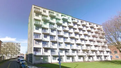 Google Maps A large white block of flats sitting on a green space, surrounded by houses and other blocks of flats. The sky is blue, and cars are parked on the road which runs to the left of the block.