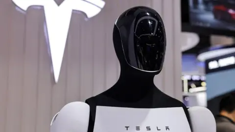 Reuters Tesla's Optimus humanoid robot stands in front of a sign displaying the company's logo.